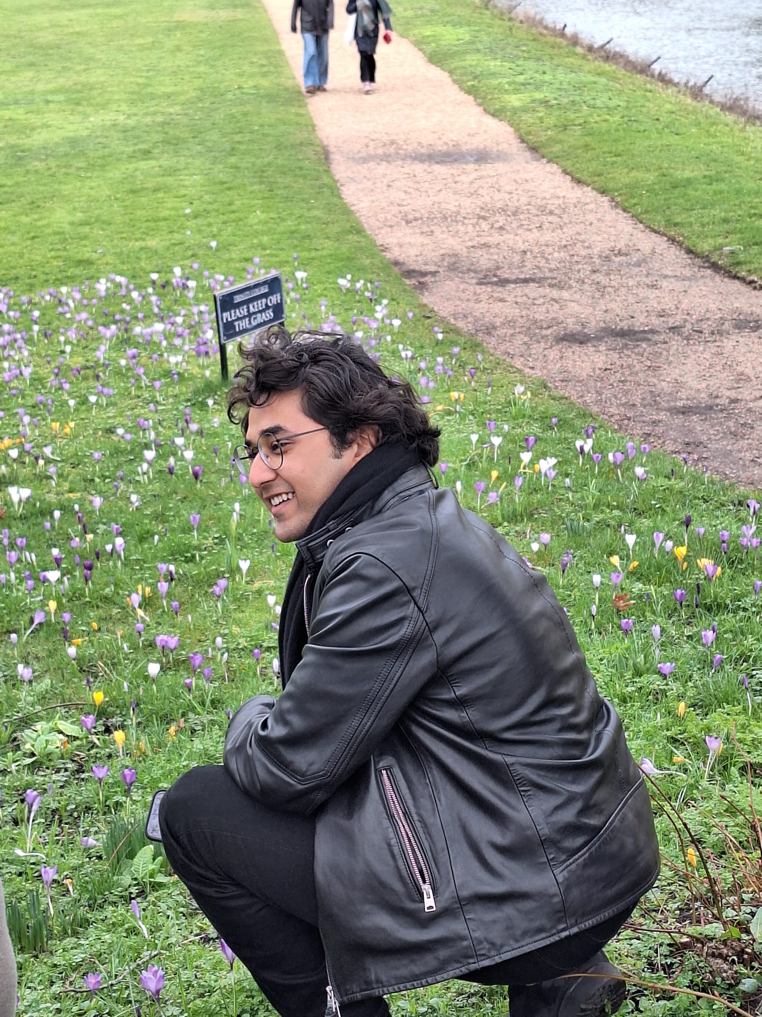 Aryan sitting on the grass next to a 'Please keep off the grass' sign, surrounded by crocuses