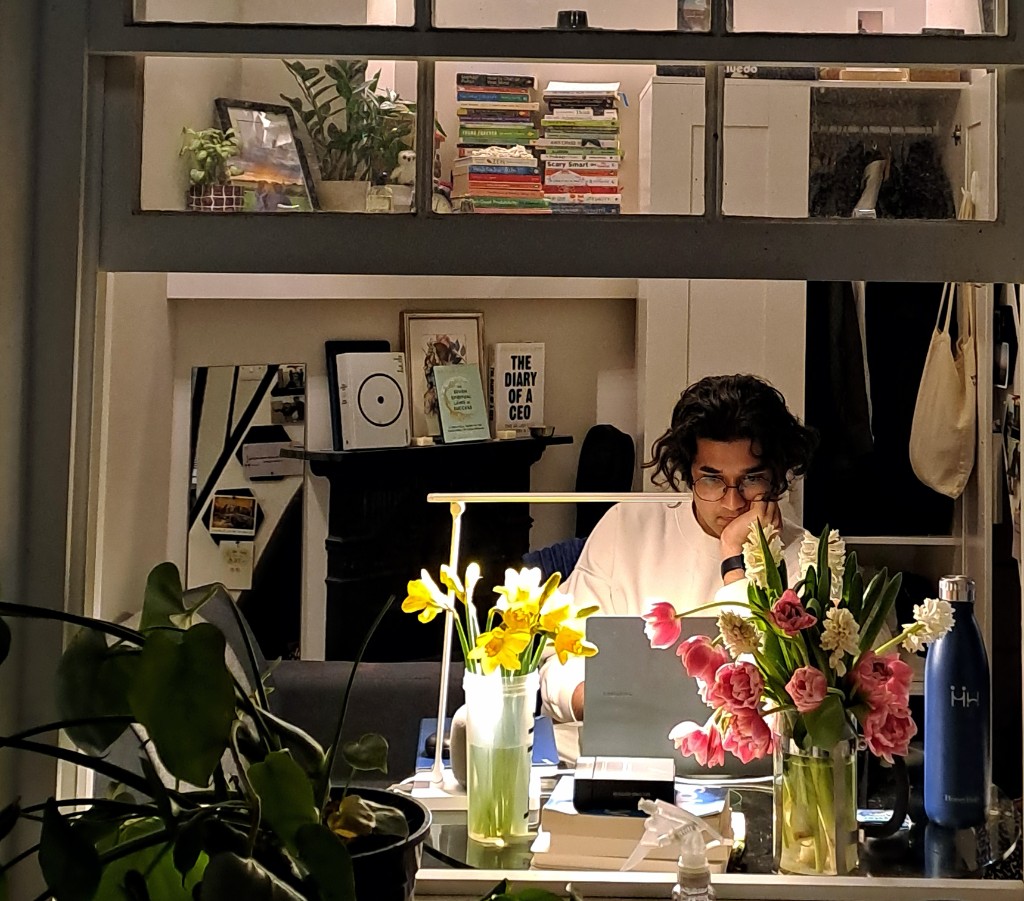 Aryan working late at a desk, surrounded by flowers and books, lamplight glowing